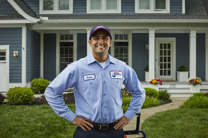 Smiling maintenance worker named Kevin in blue uniform and Roto-Rooter cap standing with hands on hips in front of a blue house with white trim.