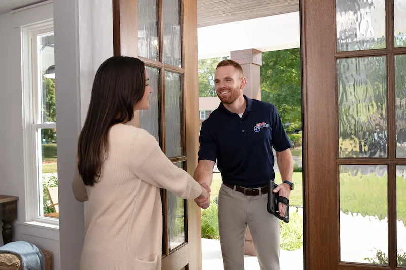 A technician in a navy blue polo shaking hands with a woman at her front door during a home service visit.