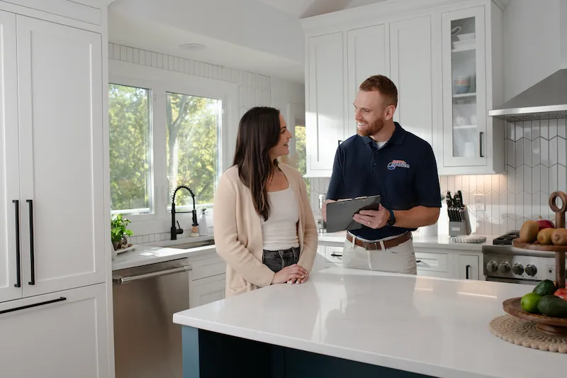A smiling man in a navy blue polo shirt holding a clipboard talks to a woman with long dark hair in a beige cardigan in a modern white kitchen.