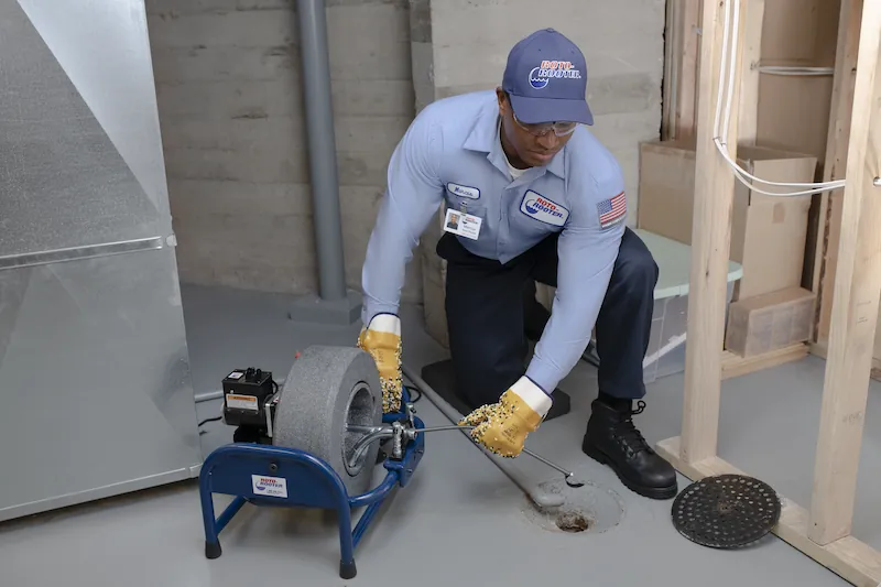 Technician in uniform using a drain cleaning machine to clear a basement floor drain.