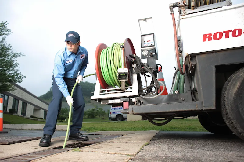 Worker in uniform handling a green hose reel connected to a ROTO truck near a drain grate outdoors.