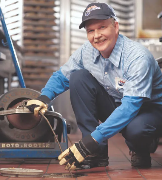 Smiling plumber in uniform using a drain cleaning machine to unclog a floor drain in a commercial kitchen.