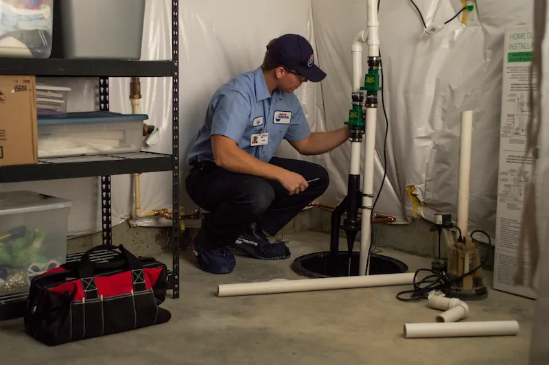 Technician in uniform inspecting and repairing a sump pump in a basement corner with tools and parts nearby.