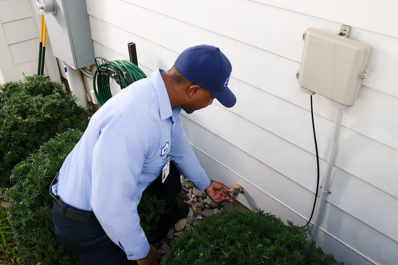 Technician in uniform crouching outdoors near a house wall, turning a garden hose faucet valve.