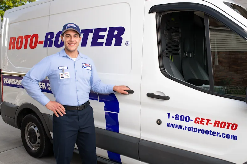A smiling Roto-Rooter technician in uniform standing next to a branded service van with the company logo and contact website.