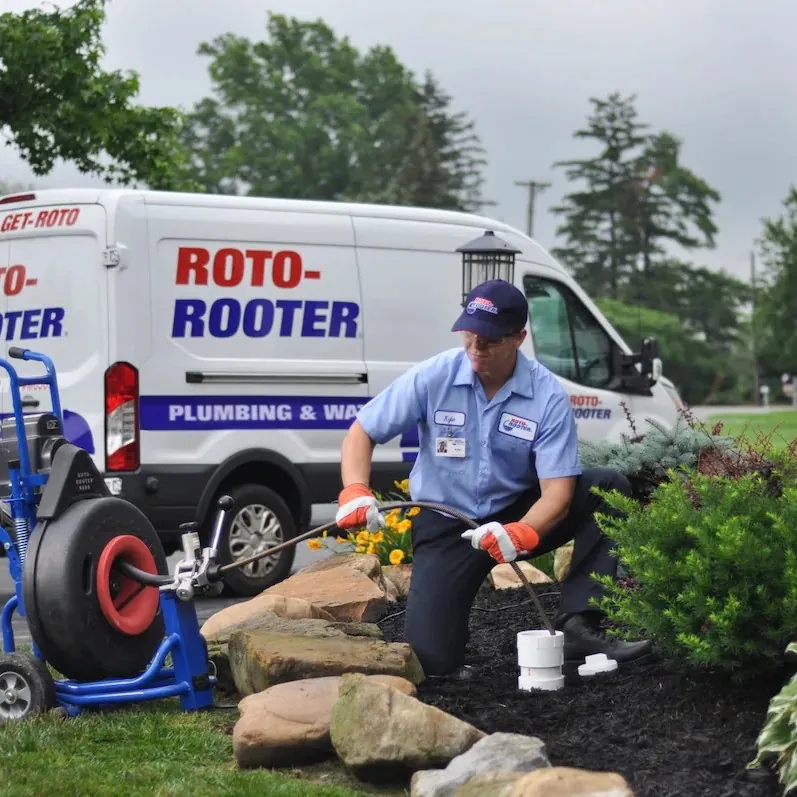 Roto-Rooter plumber using equipment to clear an underground pipe near a house with a Roto-Rooter van parked in the background.