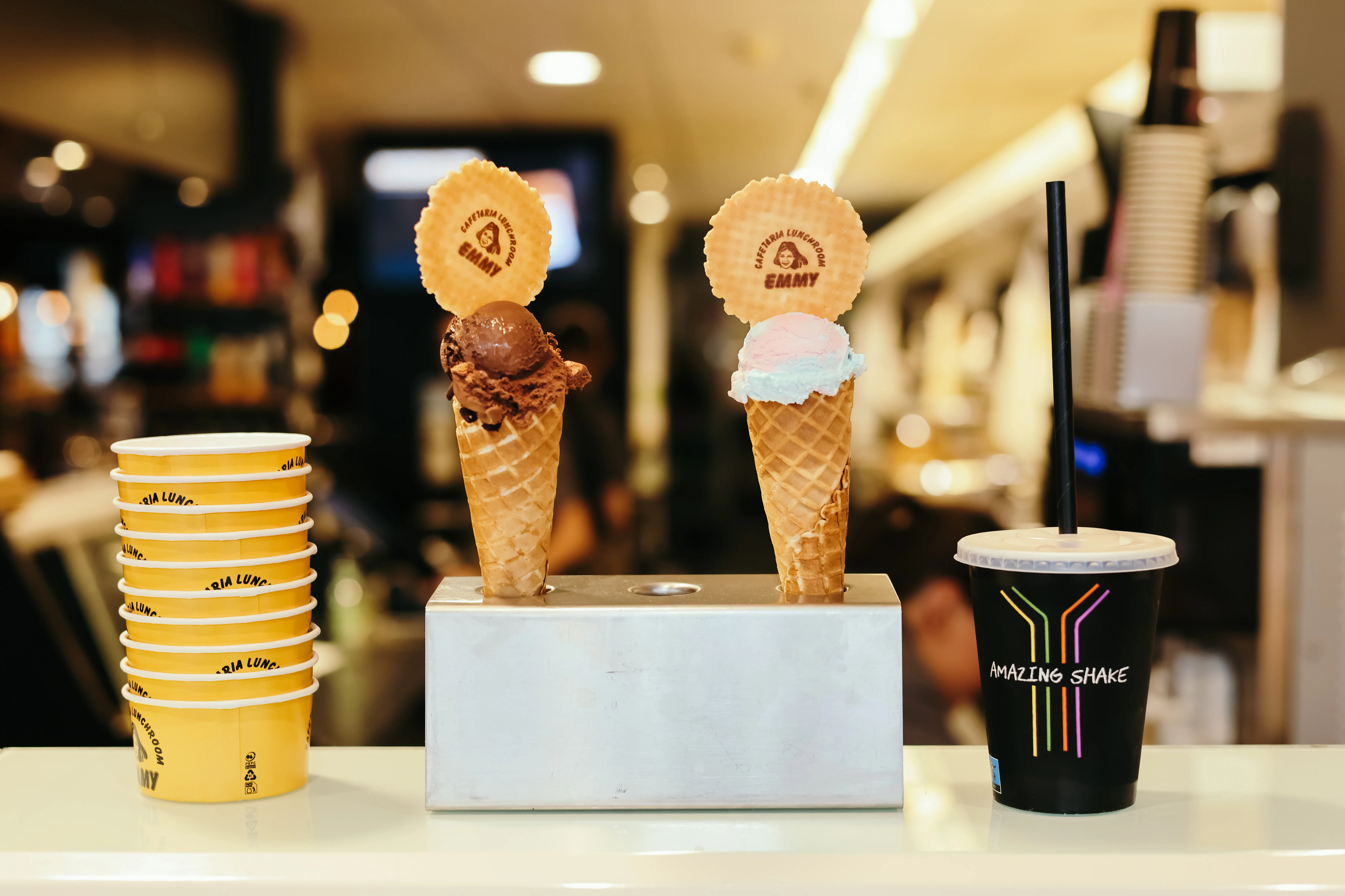 Twee ijshoorntjes, één met chocolade-ijs en één met roze en blauw ijs, elk met een wafel met 'Cafeteria Lunchroom Emmy', naast een stapel gele ijsbekers en een zwarte beker met een rietje met tekst 'Amazing Shake'.