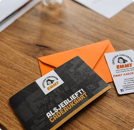 Een zwarte cadeaubon met tekst 'Alsjeblieft! Cadeaukaart' en een oranje enveloppe op een houten tafel, naast een visitekaartje van Cafetaria Lunchroom Emmy.