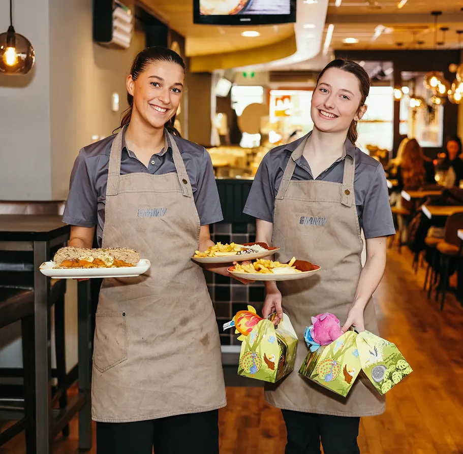 Twee glimlachende serveersters in een restaurant met bruine schorten, één draagt borden met eten, de ander draagt cadeautasjes.