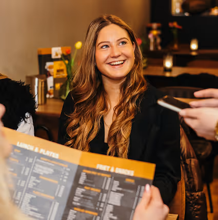 Vrouw met lang golvend haar glimlacht terwijl ze in een restaurant een menu bekijkt en een ober een apparaat aanbiedt.