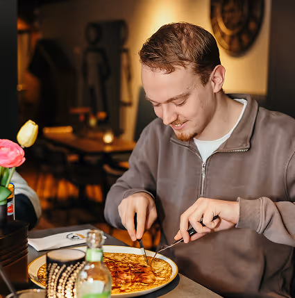 Een jongeman snijdt een pizza aan tafel in een gezellige restaurantomgeving.