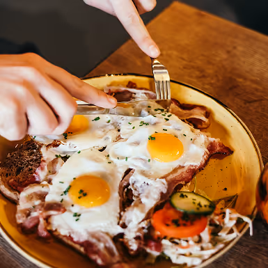 Handen die een vork en mes gebruiken om een bord met twee gebakken eieren op spek, toast en een kleine salade te eten.