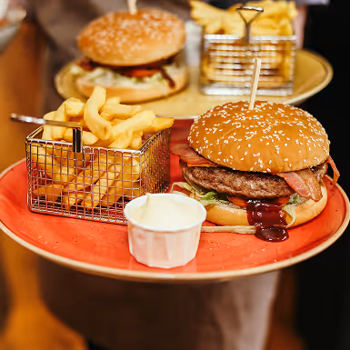 Bacon cheeseburger met sla en saus op een sesambroodje, geserveerd met frietjes in een klein metalen mandje en een bakje mayonaise op een oranje bord.