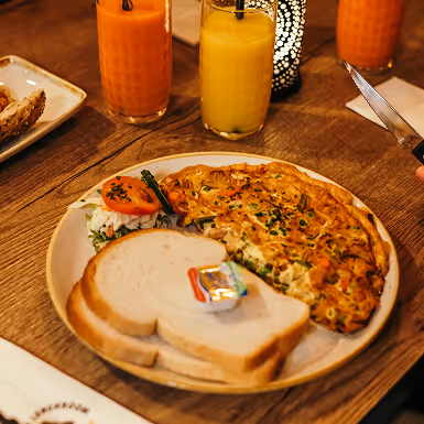 Braadpan met omelet, twee sneetjes witbrood met boter en plakje tomaat, geserveerd met sinaasappel- en wortelsap op houten tafel.