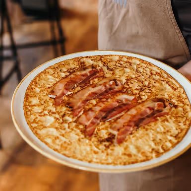Een bord met een grote, dunne pannenkoek bedekt met vijf reepjes krokant gebakken spek, vastgehouden door een persoon met een beige schort.