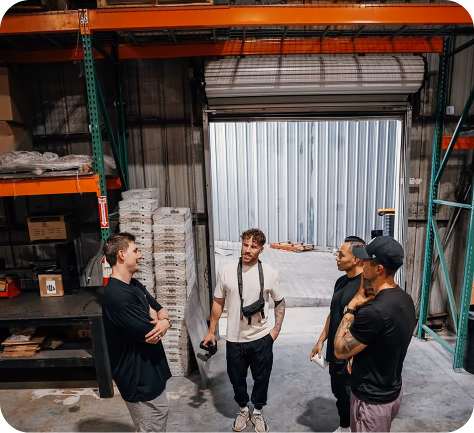 Four men standing in a warehouse having a conversation near pallets and metal shelving.