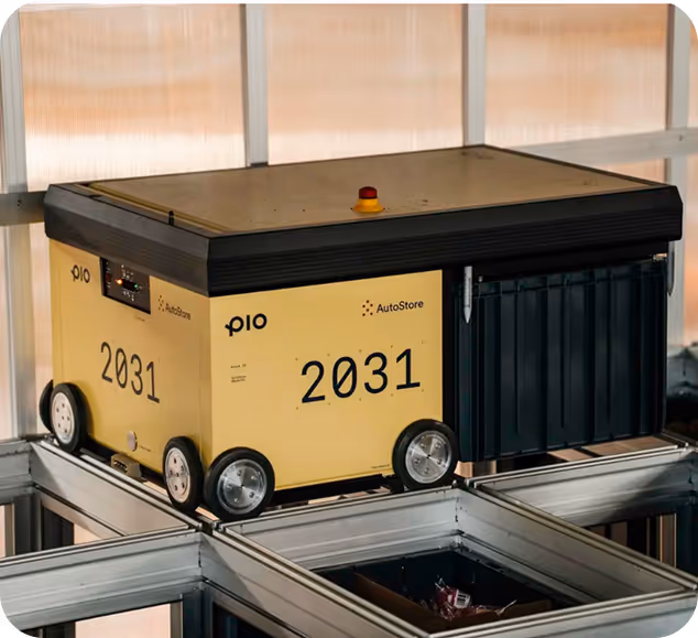 Yellow AutoStore Pio robot with number 2031 transporting a black container on a metal grid structure inside a warehouse.