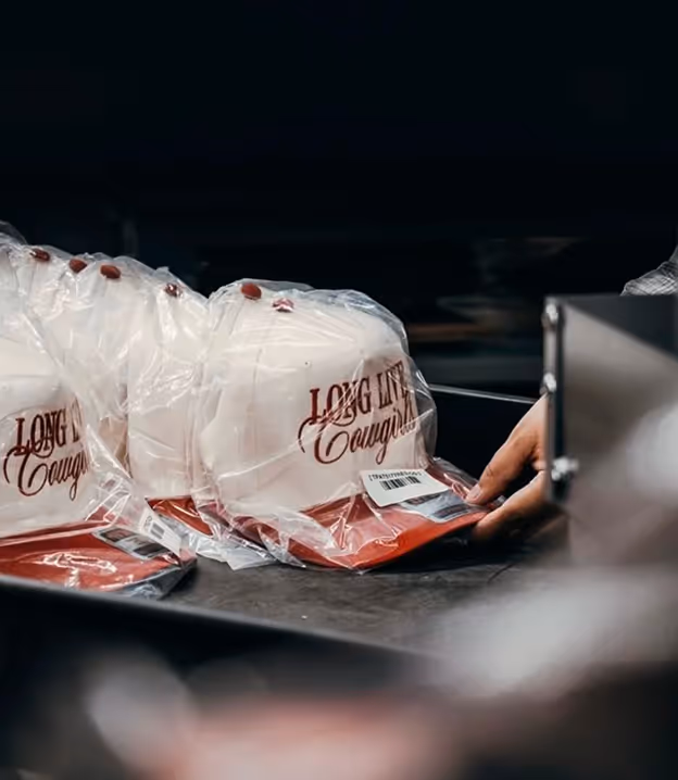 Stack of white and red caps labeled 'Long Live Cowboys' wrapped in plastic with a hand reaching towards one on a conveyor belt.