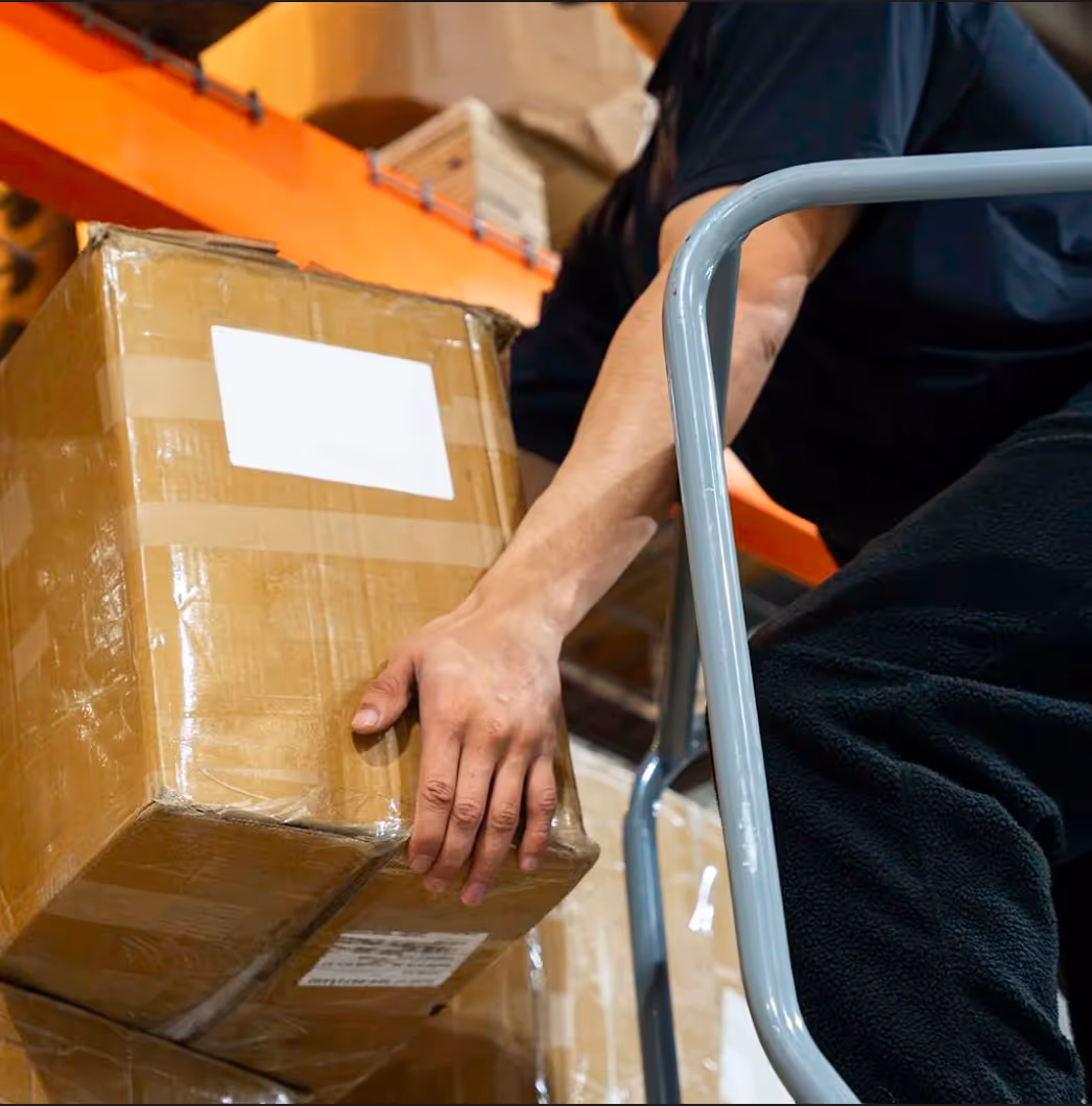 Person lifting a cardboard box in a warehouse with orange shelving in the background.