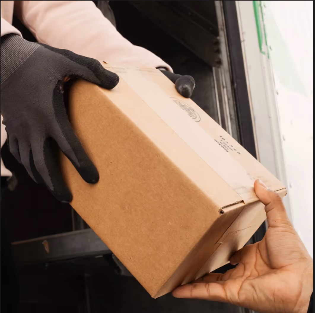 Person wearing black gloves handing a cardboard box to another person with bare hands near a truck.