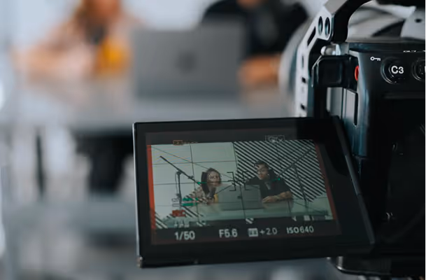 Camera screen displaying two people sitting at a table, with one person pointing at a laptop screen.