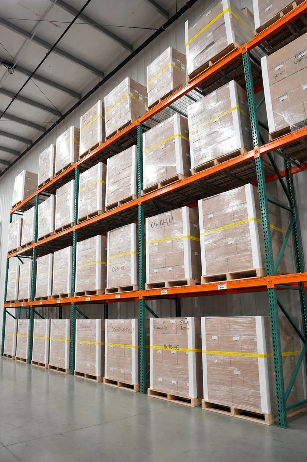 Large warehouse shelves stacked with pallets wrapped in plastic and secured by yellow straps.