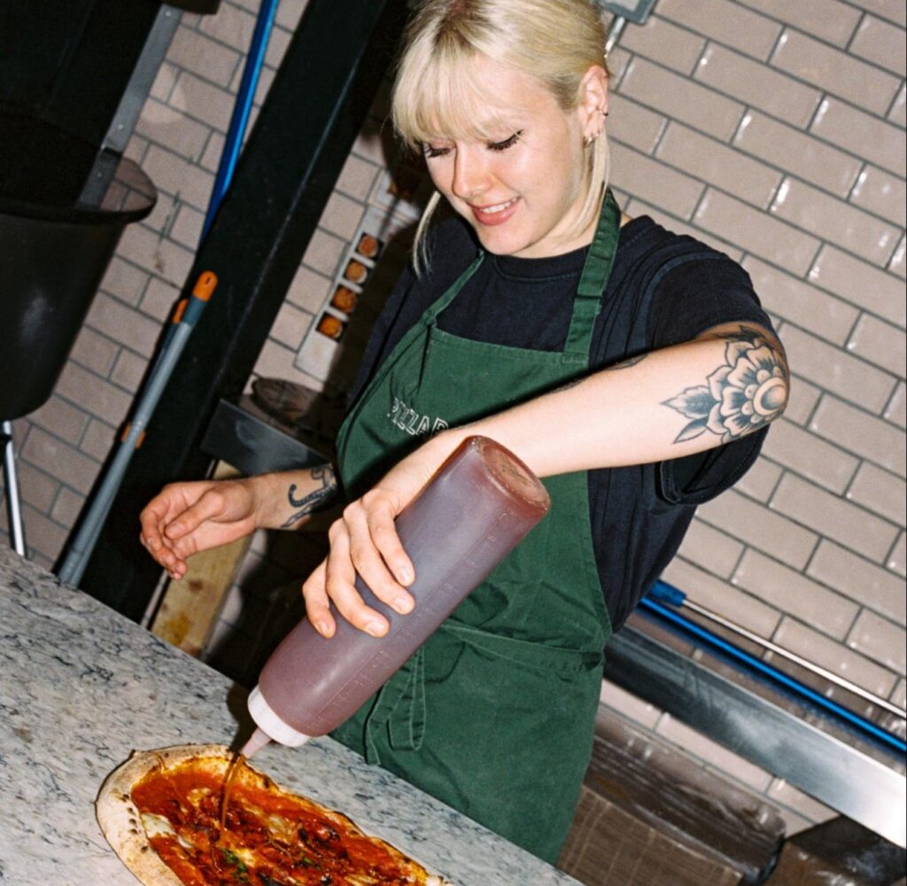 Restaurant worker adding sauce to pizza while smiling
