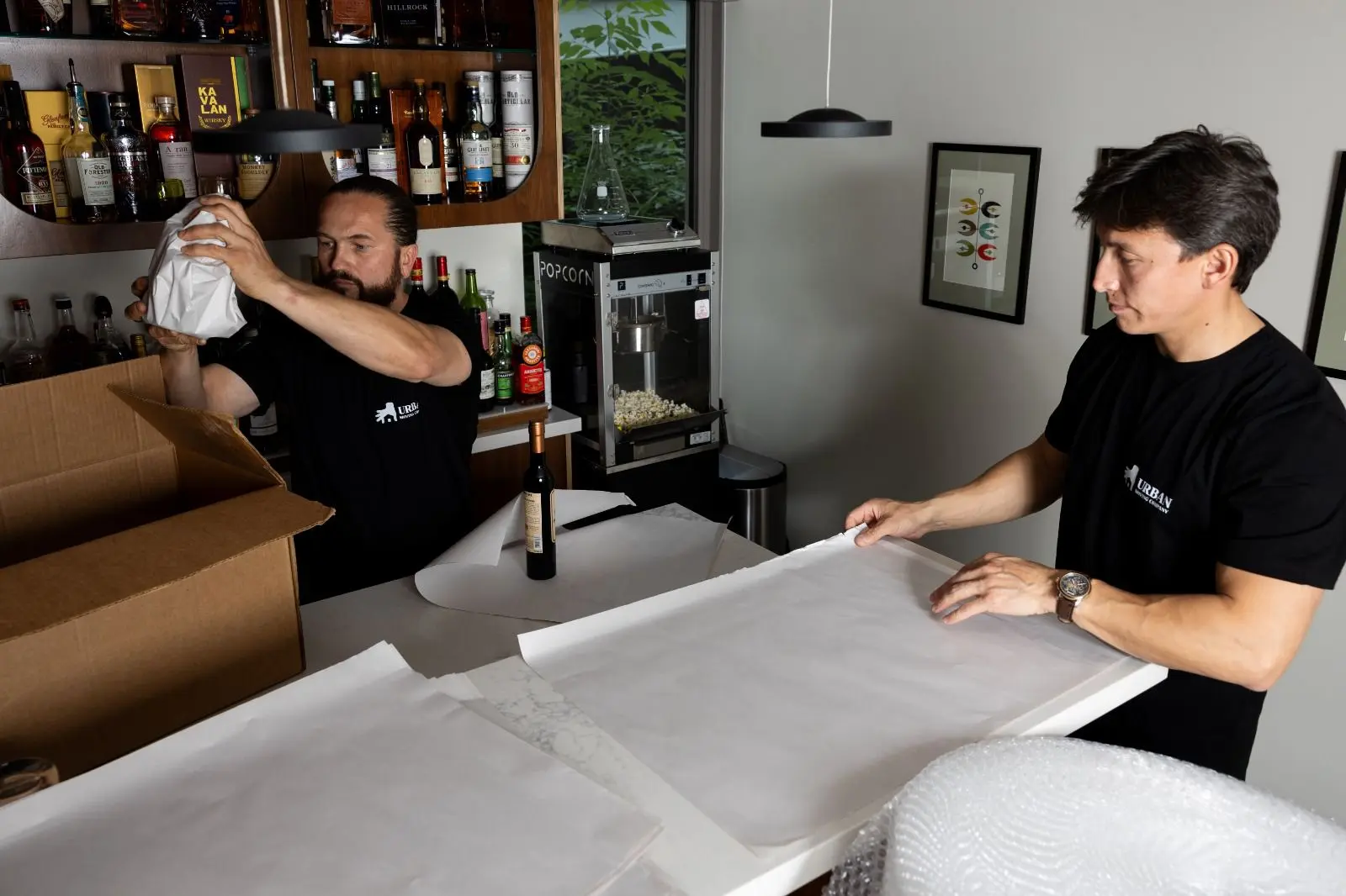 Two movers from Urban Moving in Los Angeles carefully wrap glass bottles with packing paper at a kitchen counter, preparing items for a safe residential home moving service.