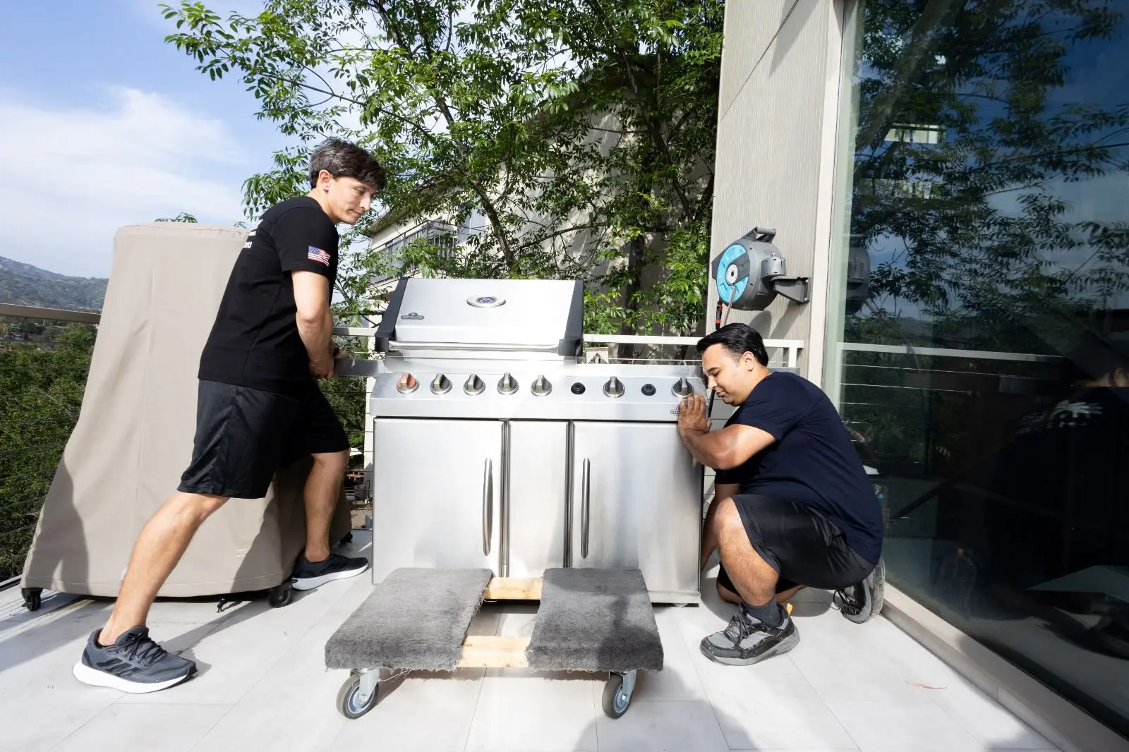 Two movers from Urban Moving in Los Angeles carefully transport a large outdoor grill using a dolly on a balcony, handling items securely for a long distance moving service.