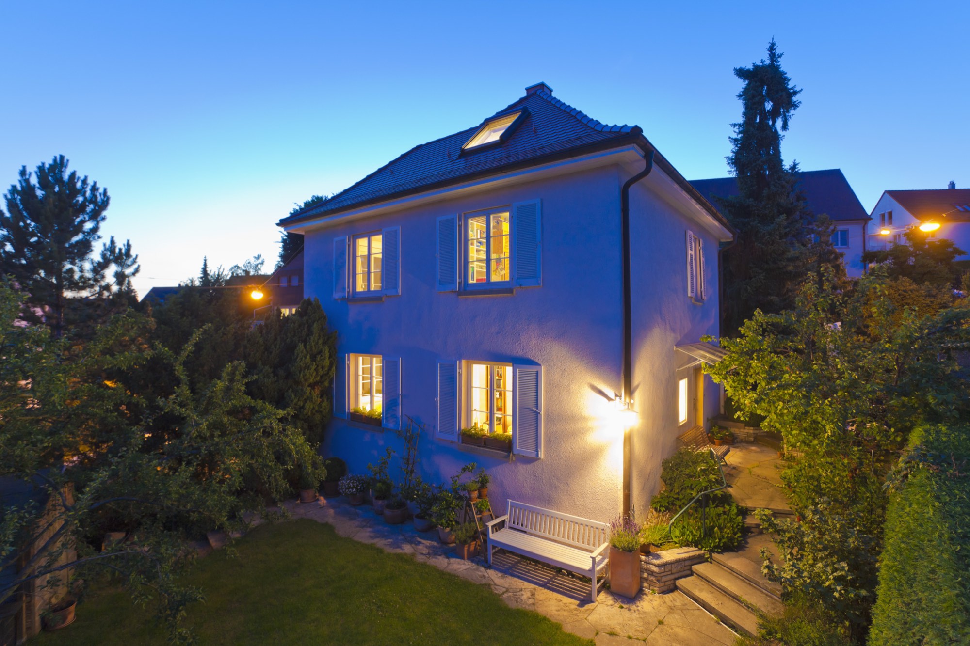 Weiß verputztes Einfamilienhaus mit Garten zur blauen Stunde, warmes Licht in den Fenstern