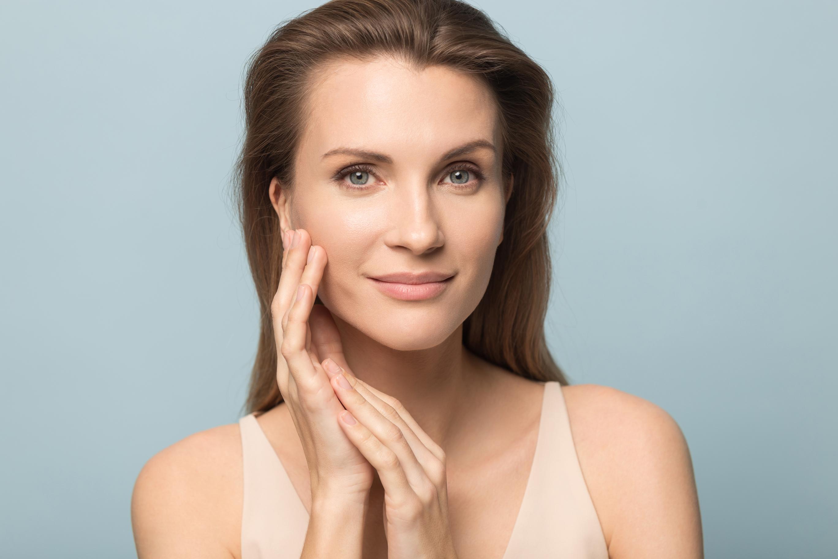 Woman with clear skin gently touching her face and smiling against a light blue background.