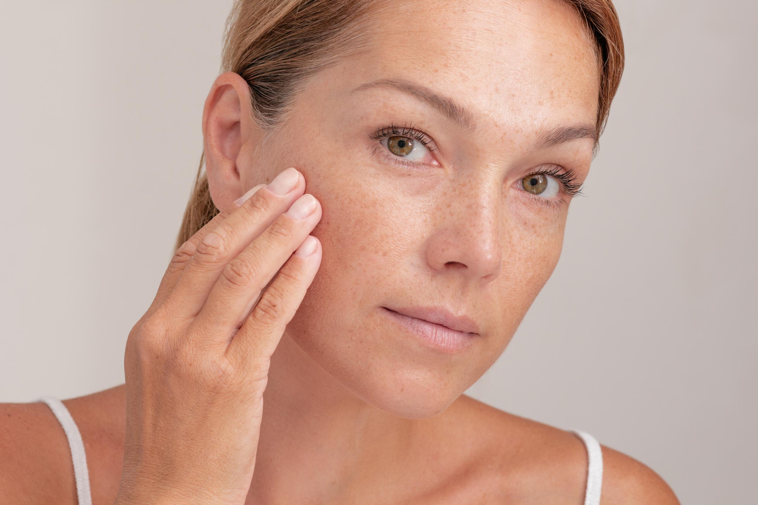 Woman with clear skin gently touching her face and smiling against a light blue background.