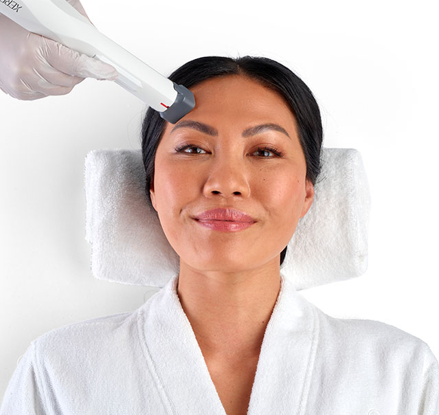 Woman with clear skin gently touching her face and smiling against a light blue background.