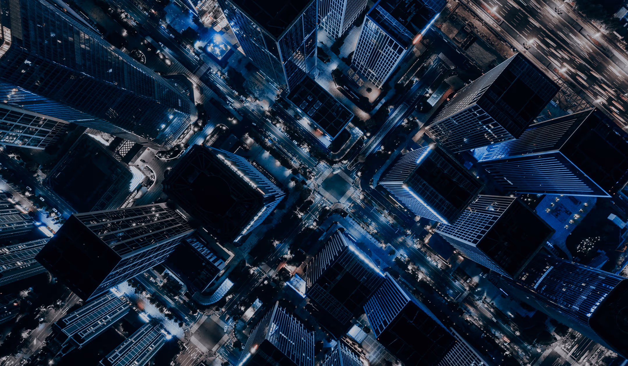 Aerial night view of a city intersection surrounded by tall illuminated skyscrapers with light trails from traffic.