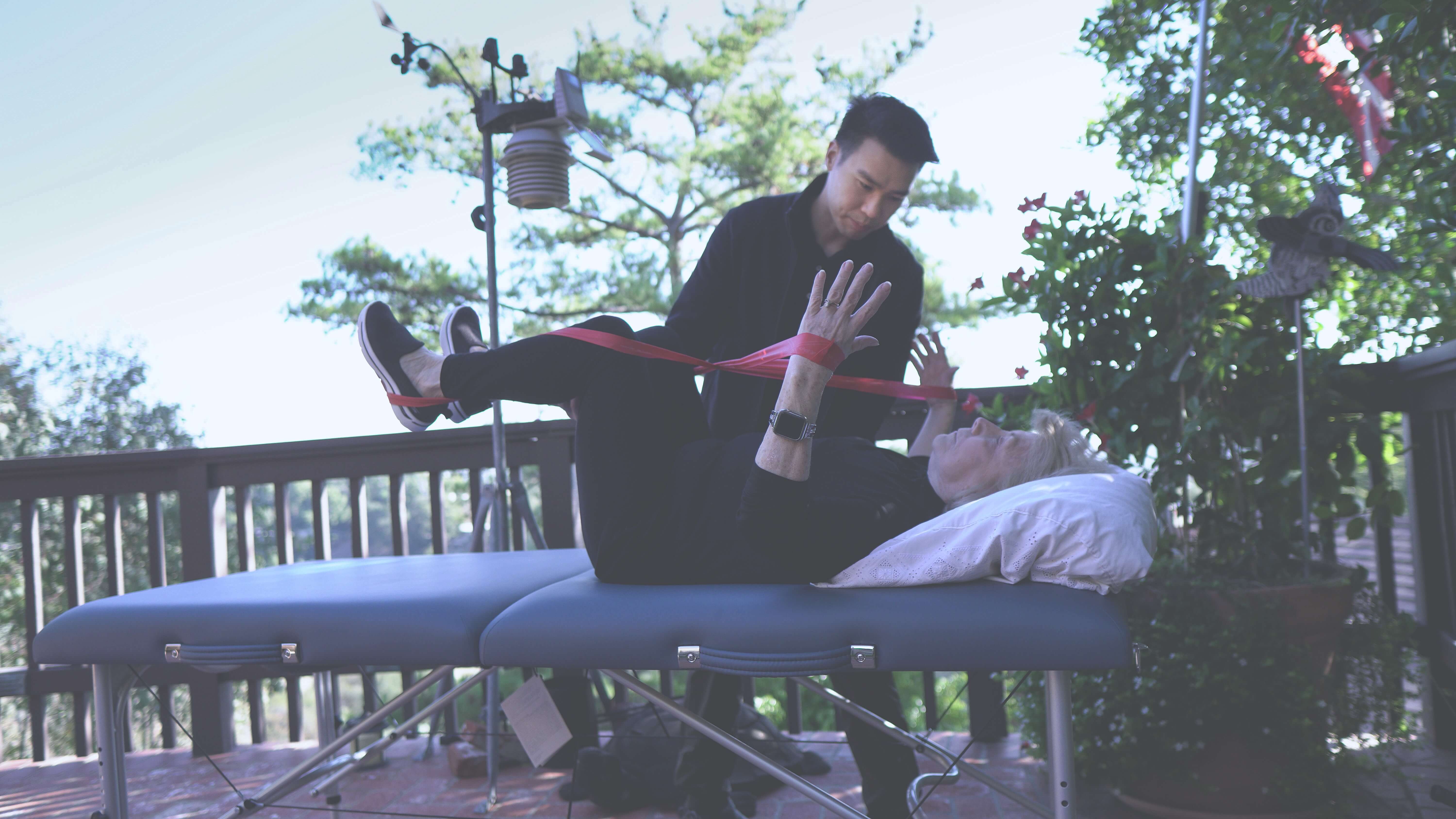 Senior woman lying on a therapy table with resistance bands on legs and arms, assisted by a man outdoors on a patio.