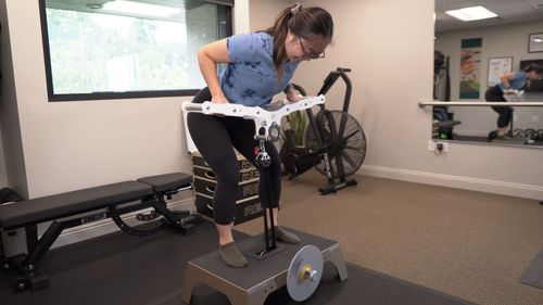 Woman in a blue shirt and black leggings performing a workout on an Exxentric kBox flywheel training machine in a gym.