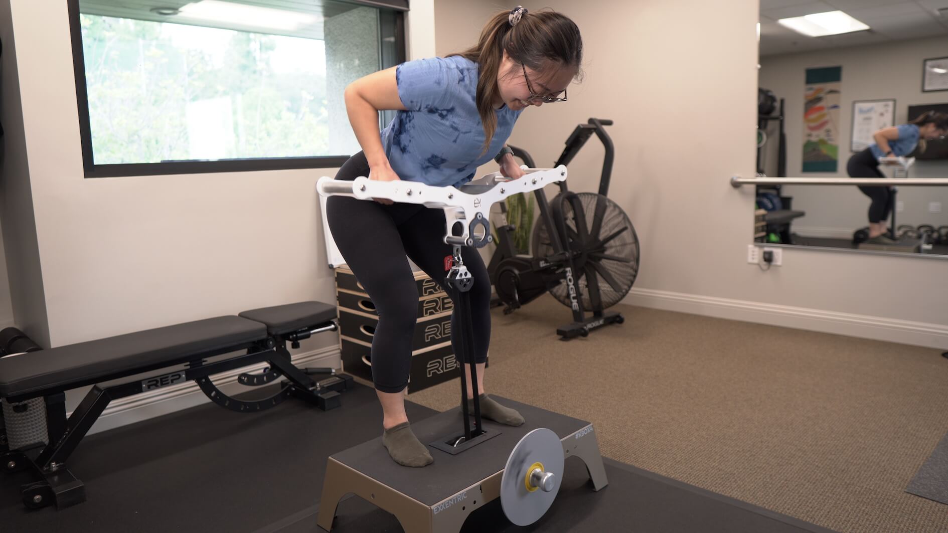 Woman in a blue shirt and black leggings performing a workout on an Exxentric kBox flywheel training machine in a gym.