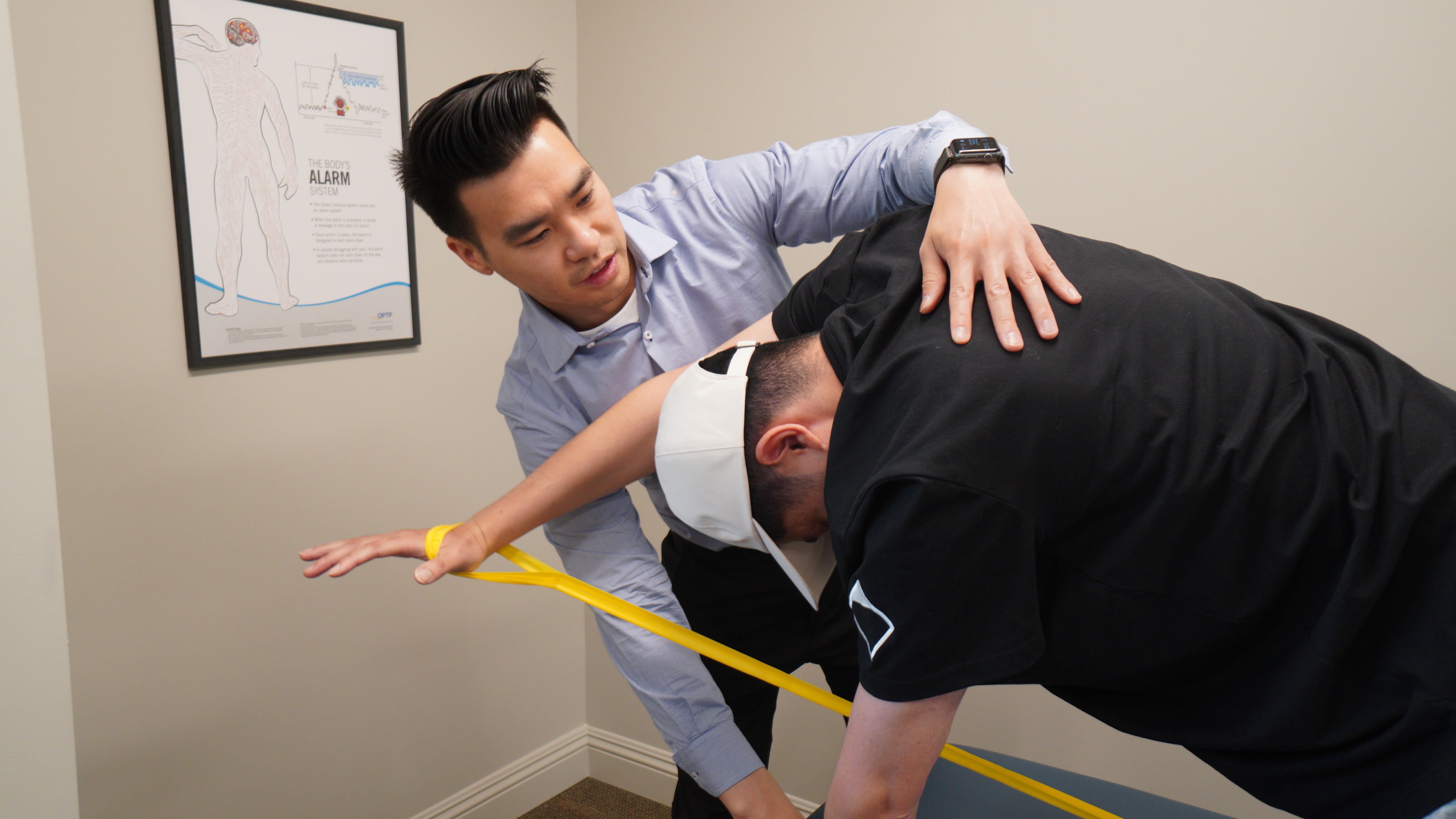 Physical therapist assisting a man wearing a white cap with a stretching exercise using a yellow resistance band in a treatment room.
