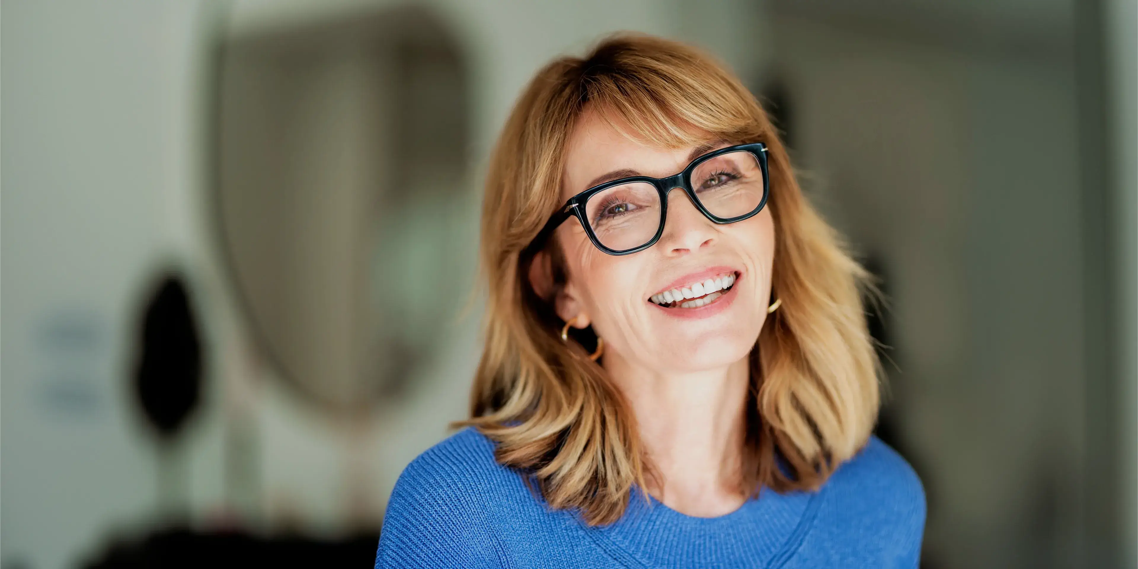 Smiling middle-aged woman with blonde hair and black glasses wearing a blue sweater in a blurred indoor setting.