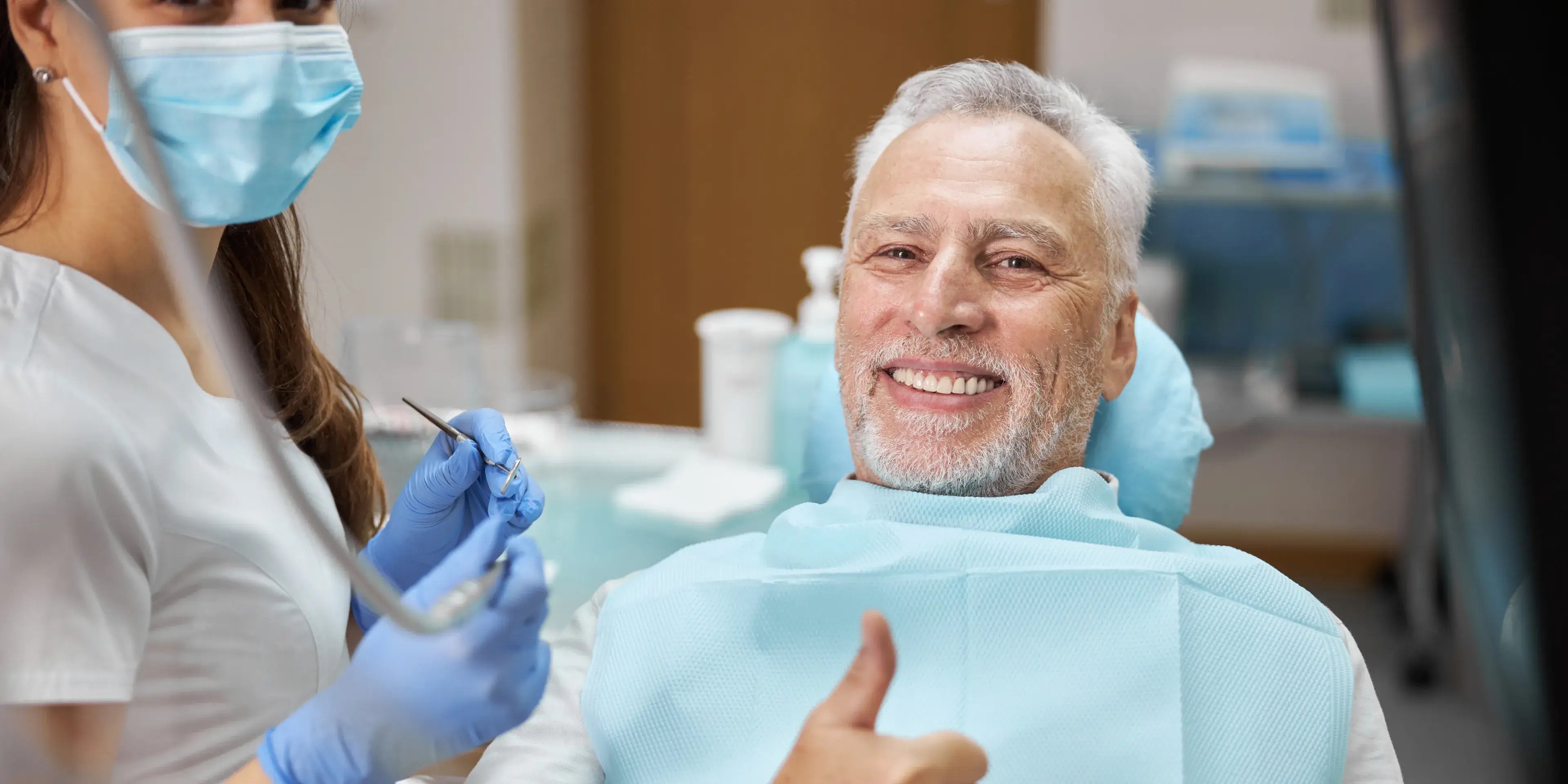 Smiling elderly man in dental chair giving thumbs up to masked dentist in gloves holding dental tools.