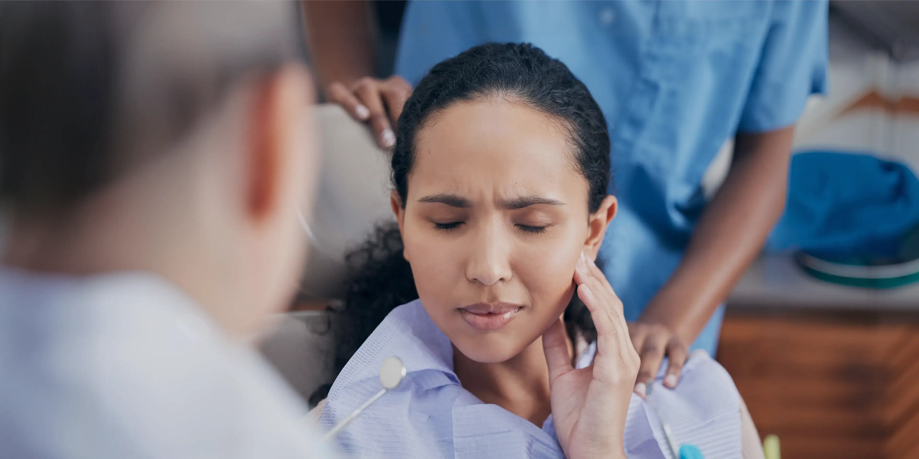 Woman in dentist chair holds her jaw with eyes closed, appearing to have dental pain.