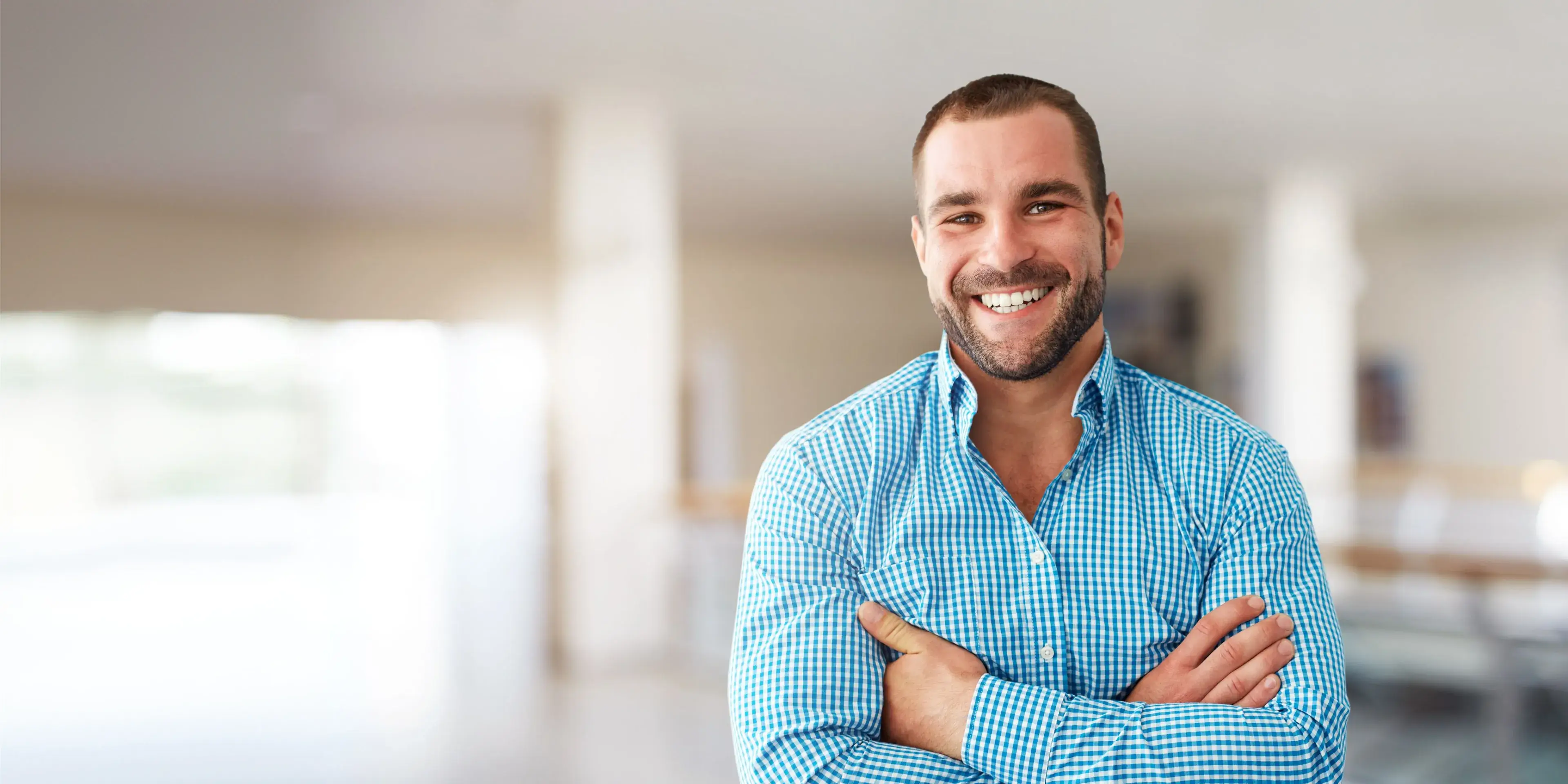 Smiling man with beard and crossed arms wearing a blue checkered shirt indoors.