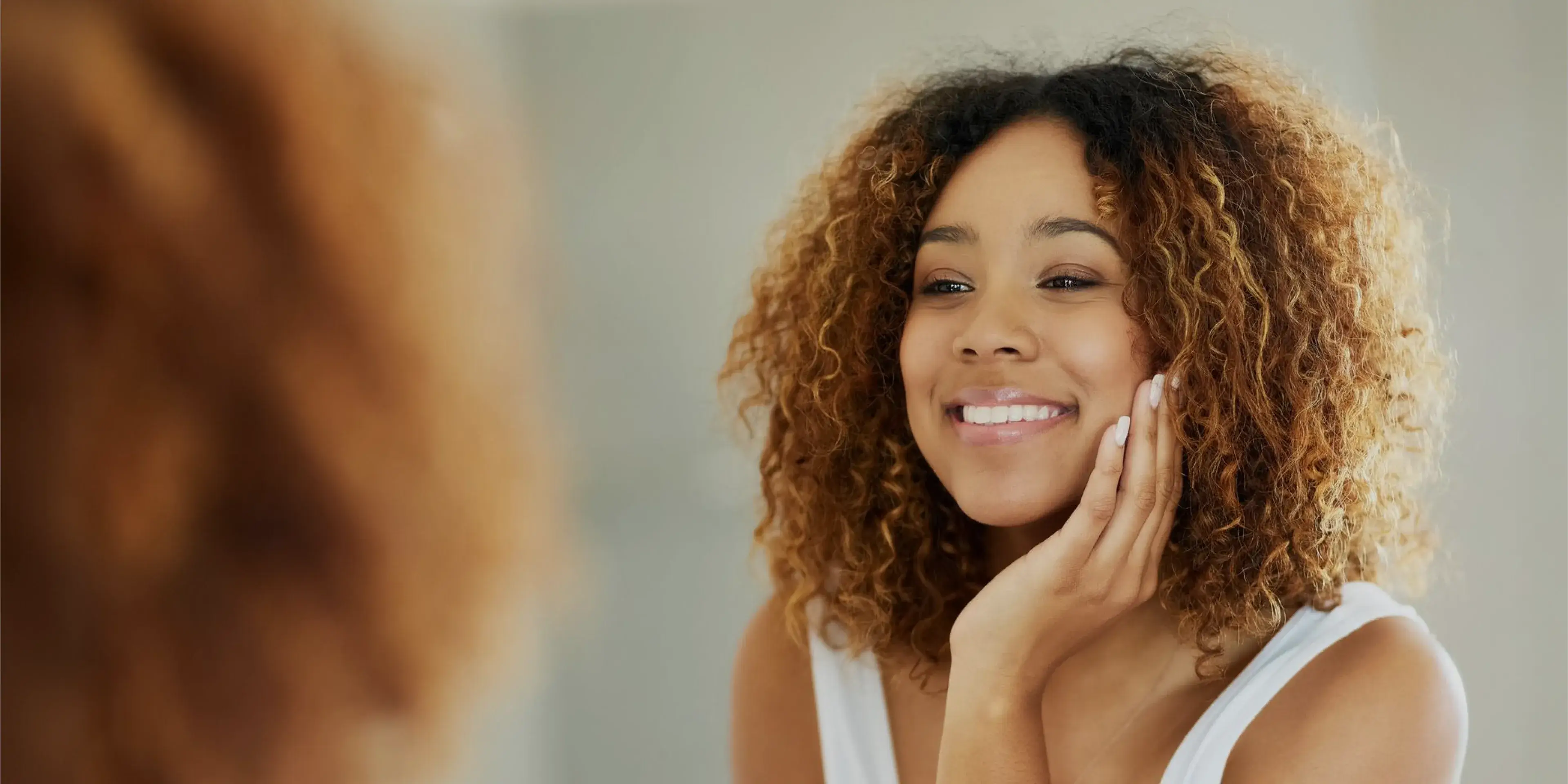 Smiling woman with curly hair touching her face while looking in a mirror.