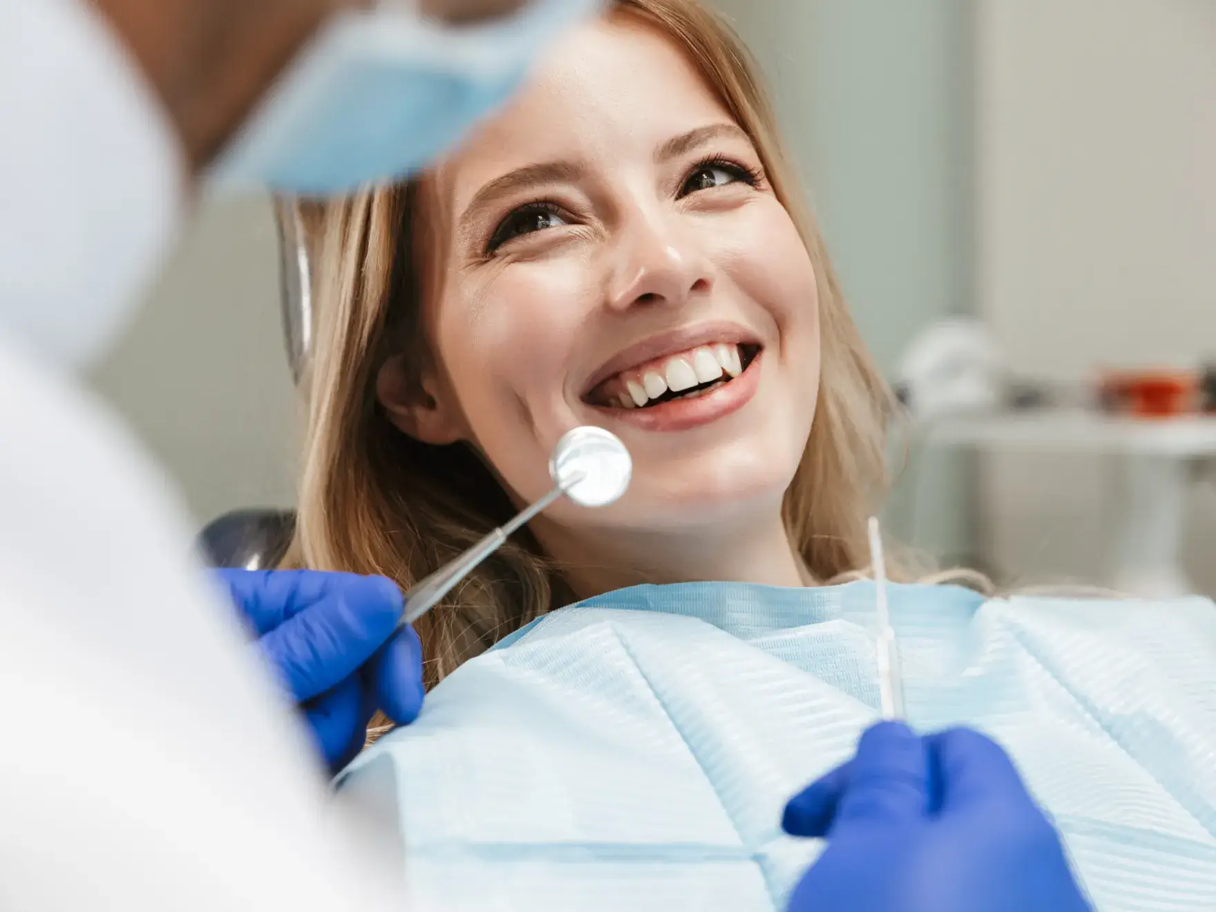 Smiling woman in dental chair as dentist wearing gloves holds dental mirror and tool.