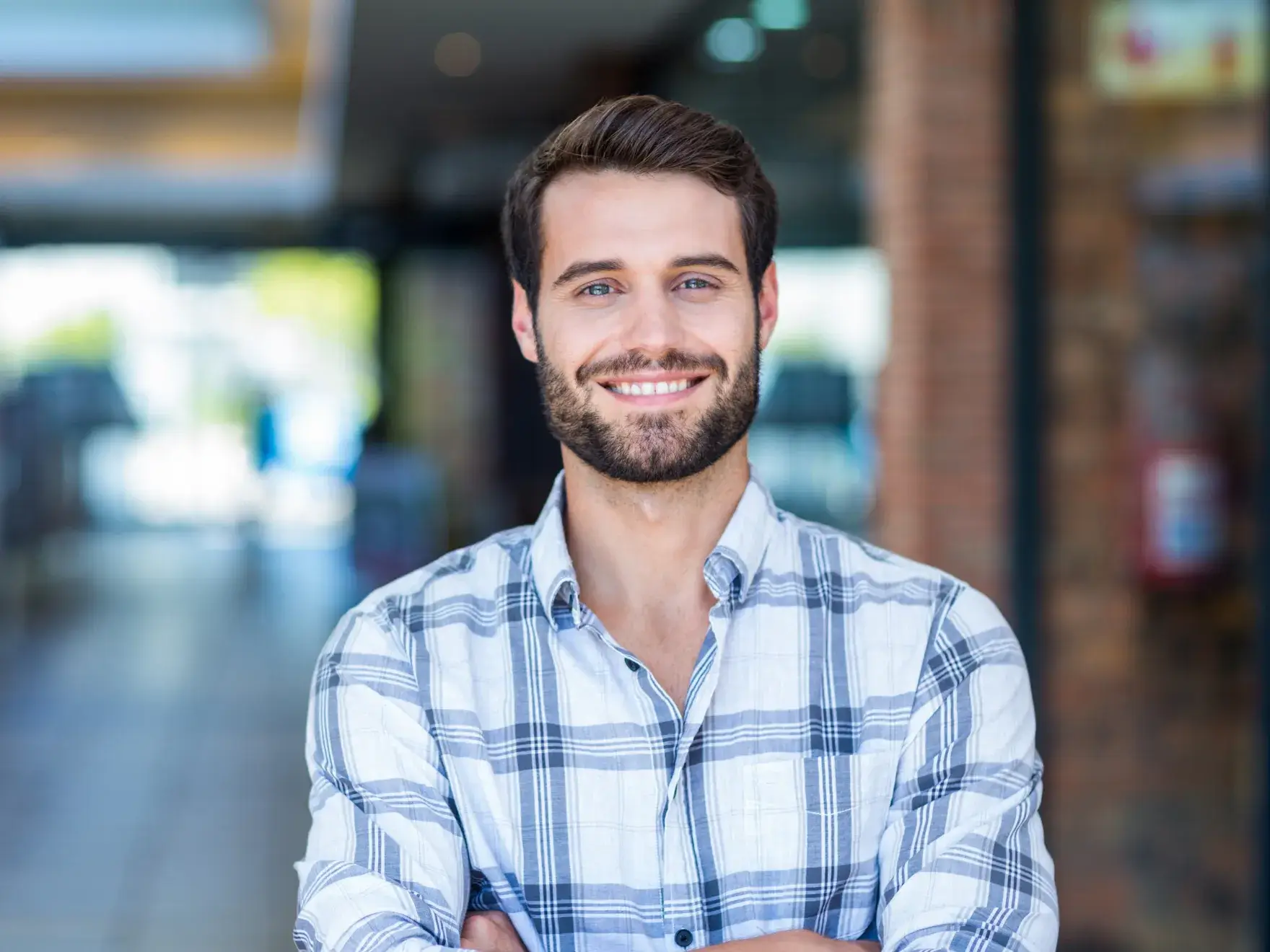 Smiling man with beard wearing a plaid shirt standing with arms crossed indoors.