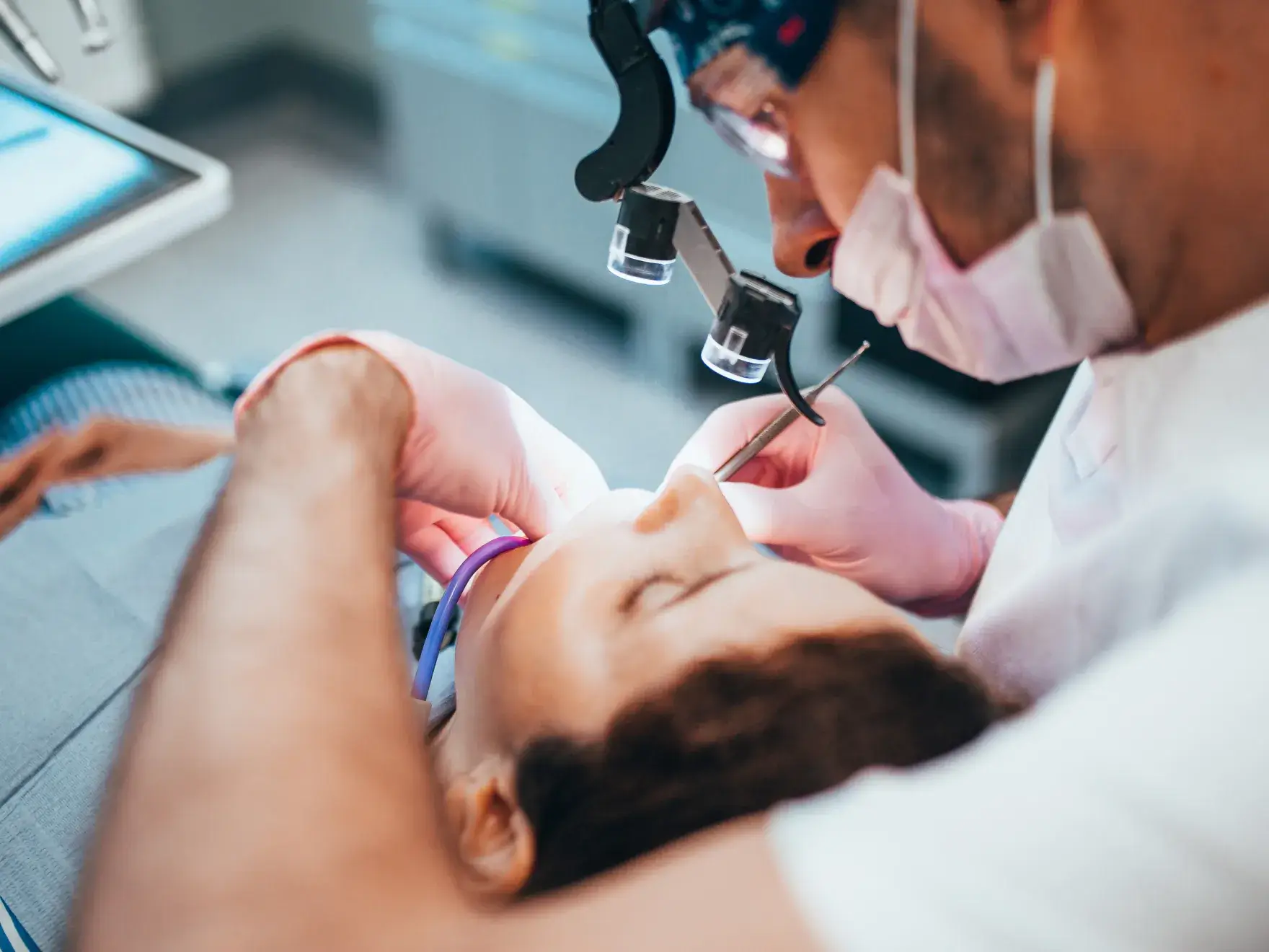 Dentist wearing magnifying glasses and mask treating a patient lying back in a dental chair.