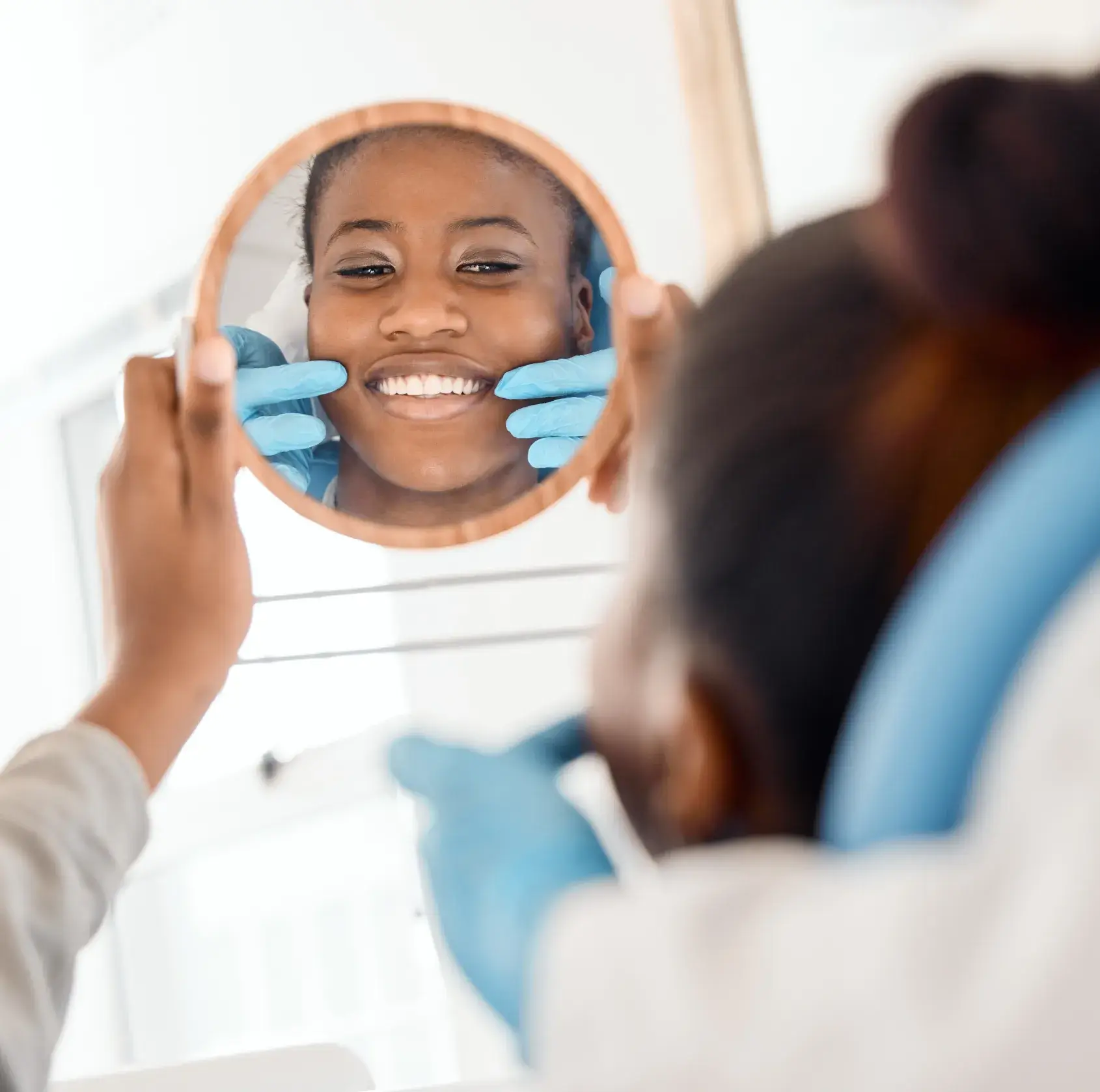 Person holding round mirror reflecting a smiling face with blue-gloved hands on cheeks.