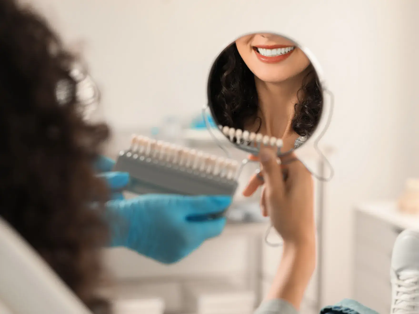 Smiling woman sees her teeth in a hand mirror while a dental professional holds a tooth shade guide.