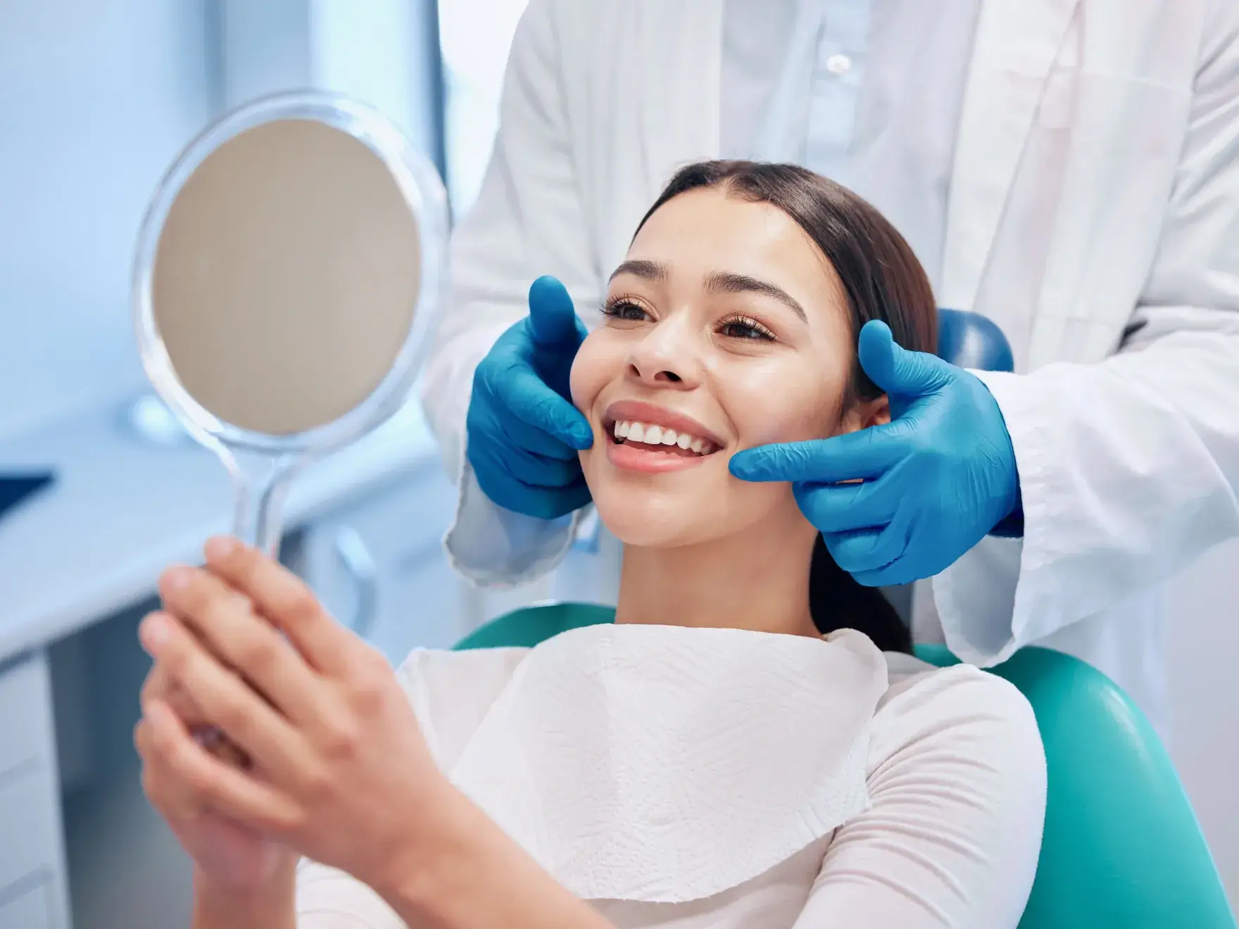 Smiling woman in dental chair holding mirror while dentist wearing gloves examines her teeth.