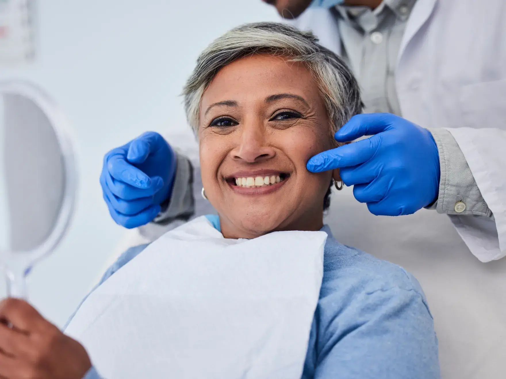 Smiling woman in a dental chair holding a mirror while dentist wearing blue gloves points at her cheek.